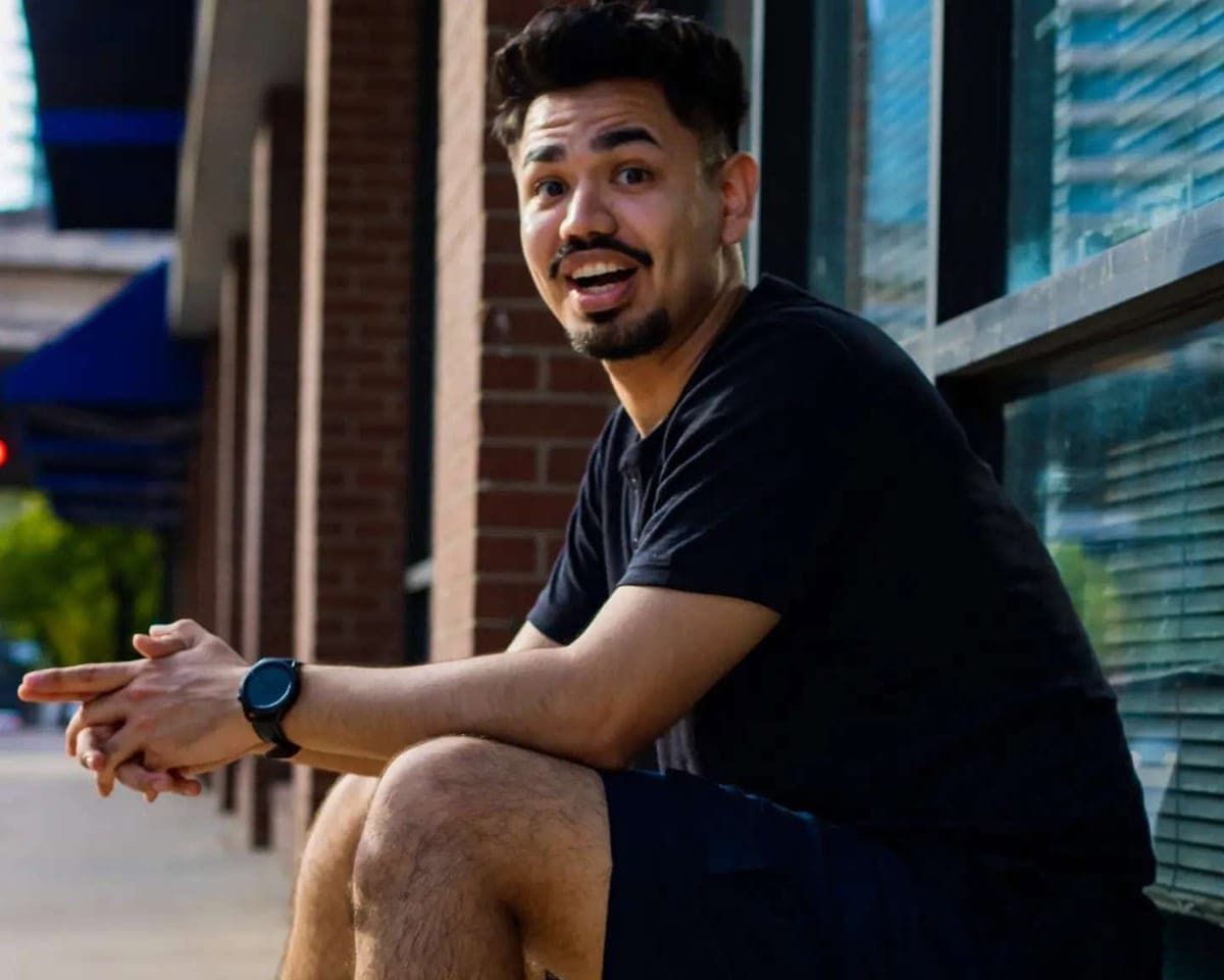Luis Gomez sitting on a brick ledge outside a building, turned toward the camera mid-laugh.