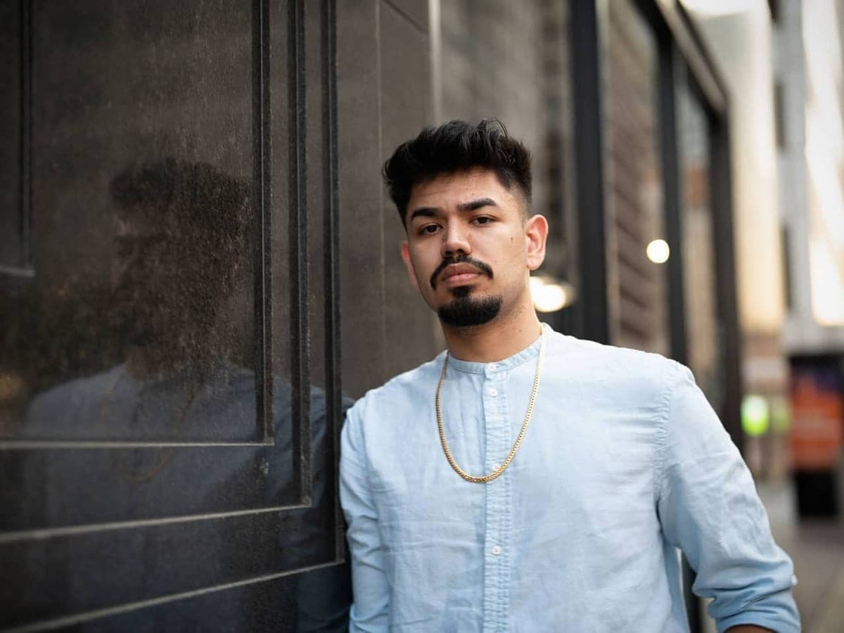 Luis Gomez leaning against a dark glass wall, his reflection visible in the pane beside him.